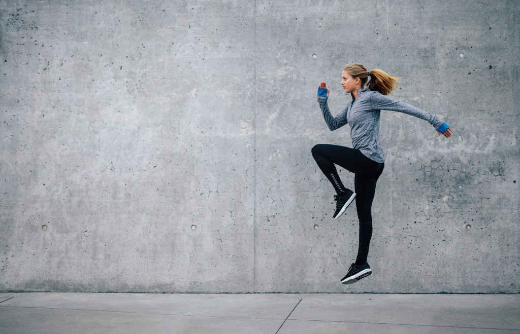 Mulher saltando em frente a uma parede cinza com roupas de ginástica