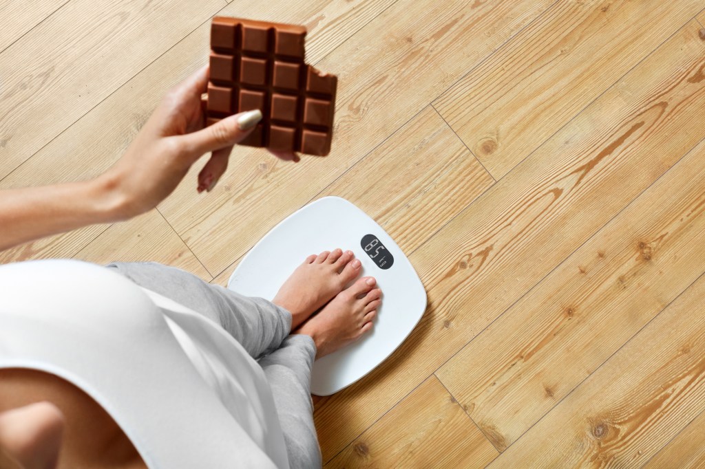 Mulher segurando uma barra de chocolate em cima de uma balança pra medir o peso