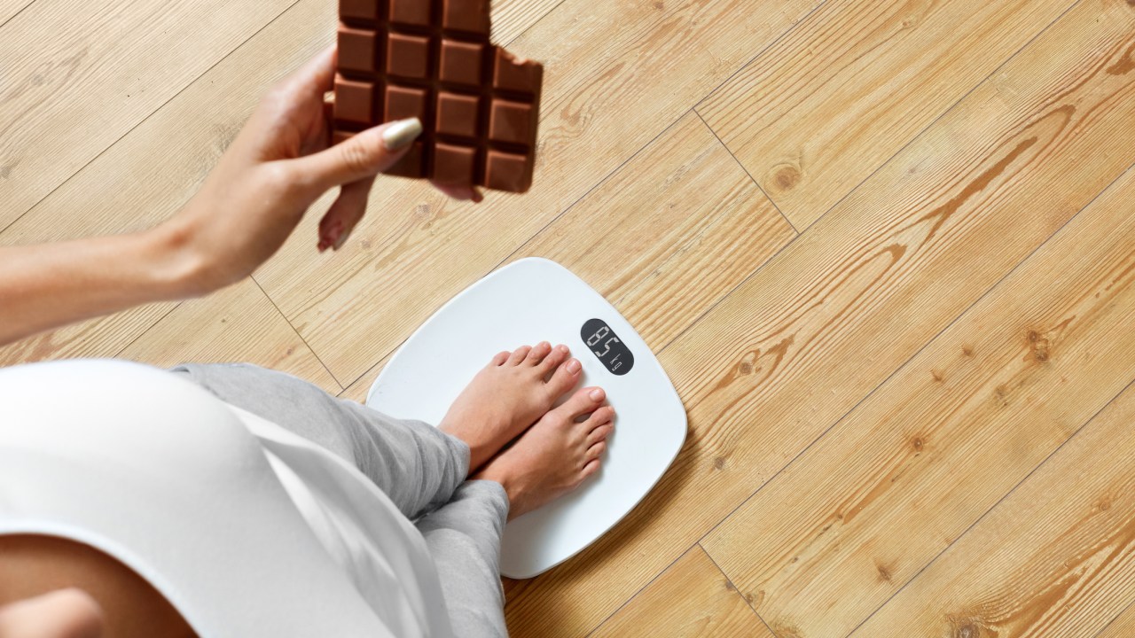 Mulher segurando uma barra de chocolate em cima de uma balança pra medir o peso