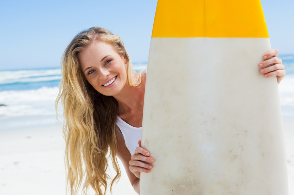 Mulher loira na praia segurando uma prancha de surfe