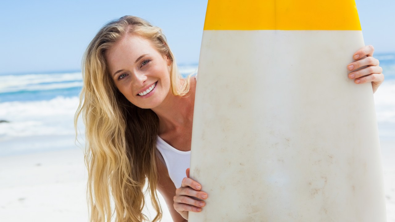 Mulher loira na praia segurando uma prancha de surfe