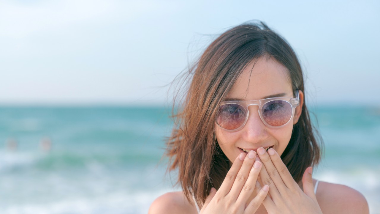 Mulher de cabelo curto na praia com as mãos na boca com cara de surpresa