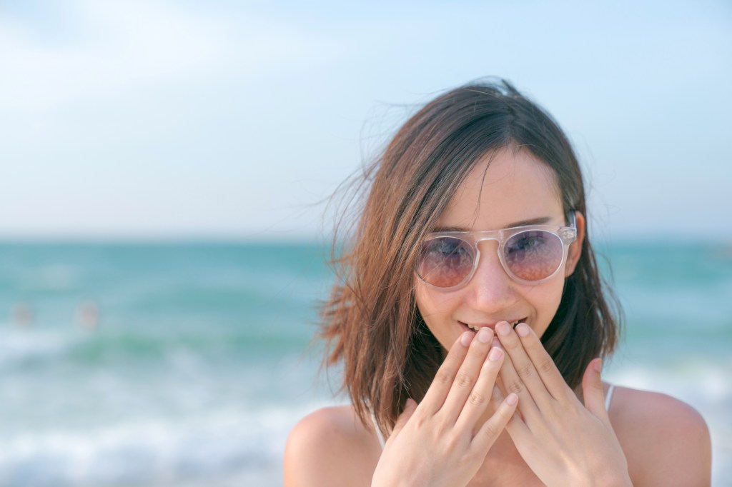 Mulher de cabelo curto na praia com as mãos na boca com cara de surpresa