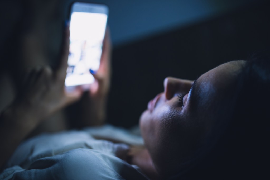 Mulher deitada na cama com a luz apagada e mexendo no celular