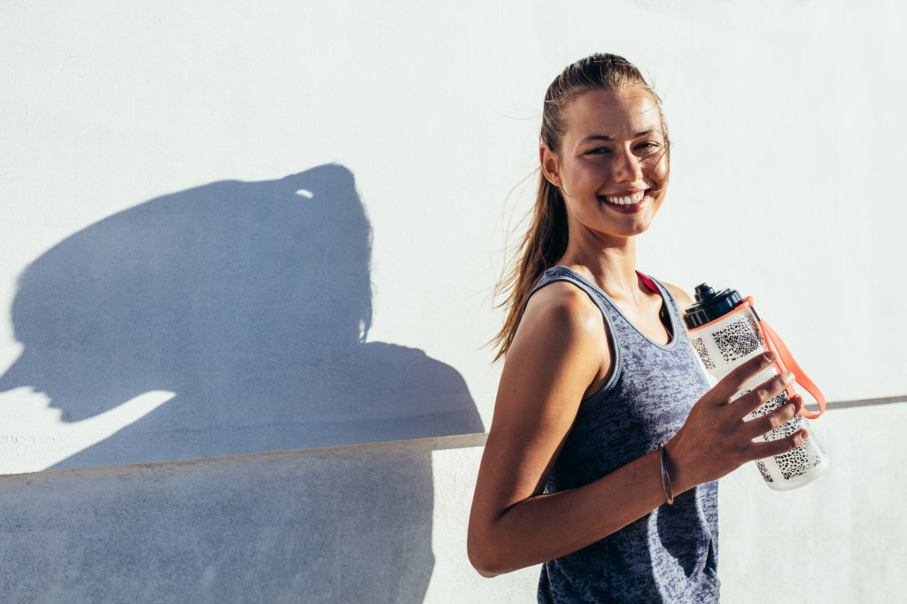 Mulher com roupa fitness fotografada numa parede clara segurando a garrafinha de água - alimentos para evitar cãibra