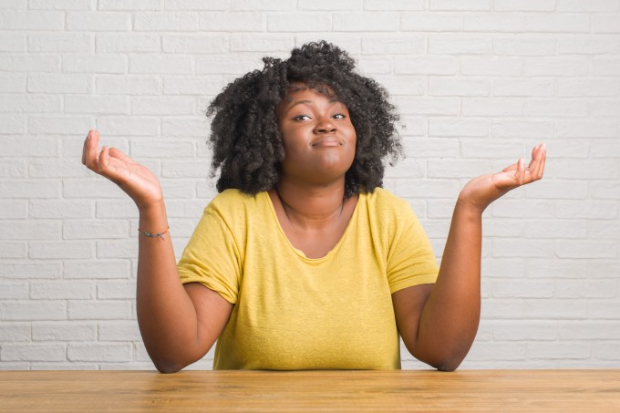 Young african american woman sitting on the table at home clueless and confused expression with arms and hands raised. Doubt concept.