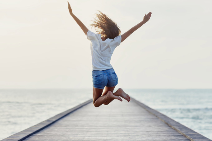 Imagem ilustrativa mostra uma mulher pulando, feliz, de frente para o mar.