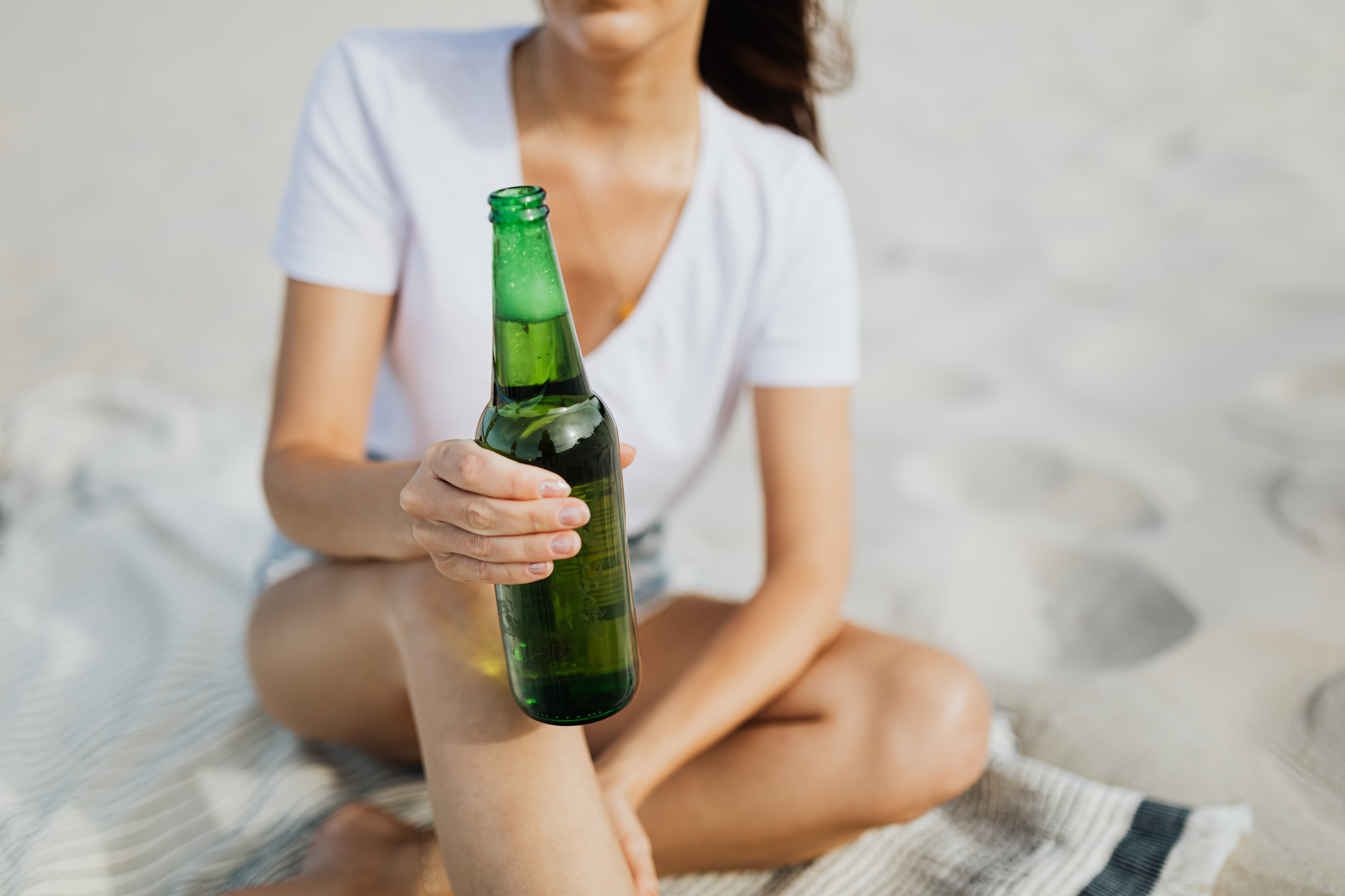 Imagem meramente ilustrativa mostra uma mulher sentada em uma toalha na praia segurando uma cerveja.