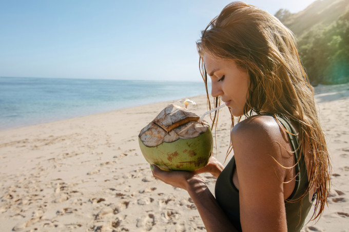 Foto ilustrativa de uma mulheres tomando água de coco na praia