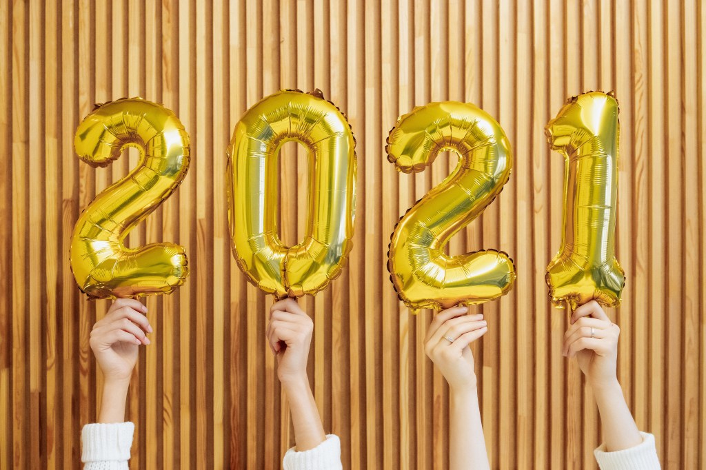 Balões dourados formando 2021, cada um sendo segurado por mãos brancas em um fundo de madeira clara