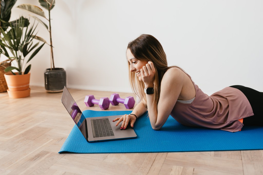 Mulher deitada em um tapete de Yoga olhando o computador ao lado de pesos para se exercitar.
