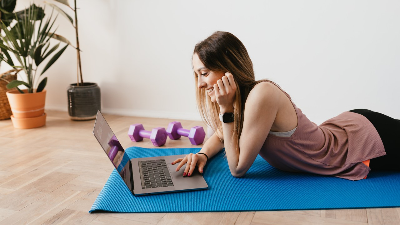 Mulher deitada em um tapete de Yoga olhando o computador ao lado de pesos para se exercitar.