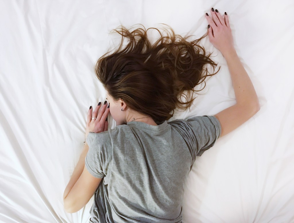 garota de camiseta cinza dormindo de bruços em cama grande e branca
