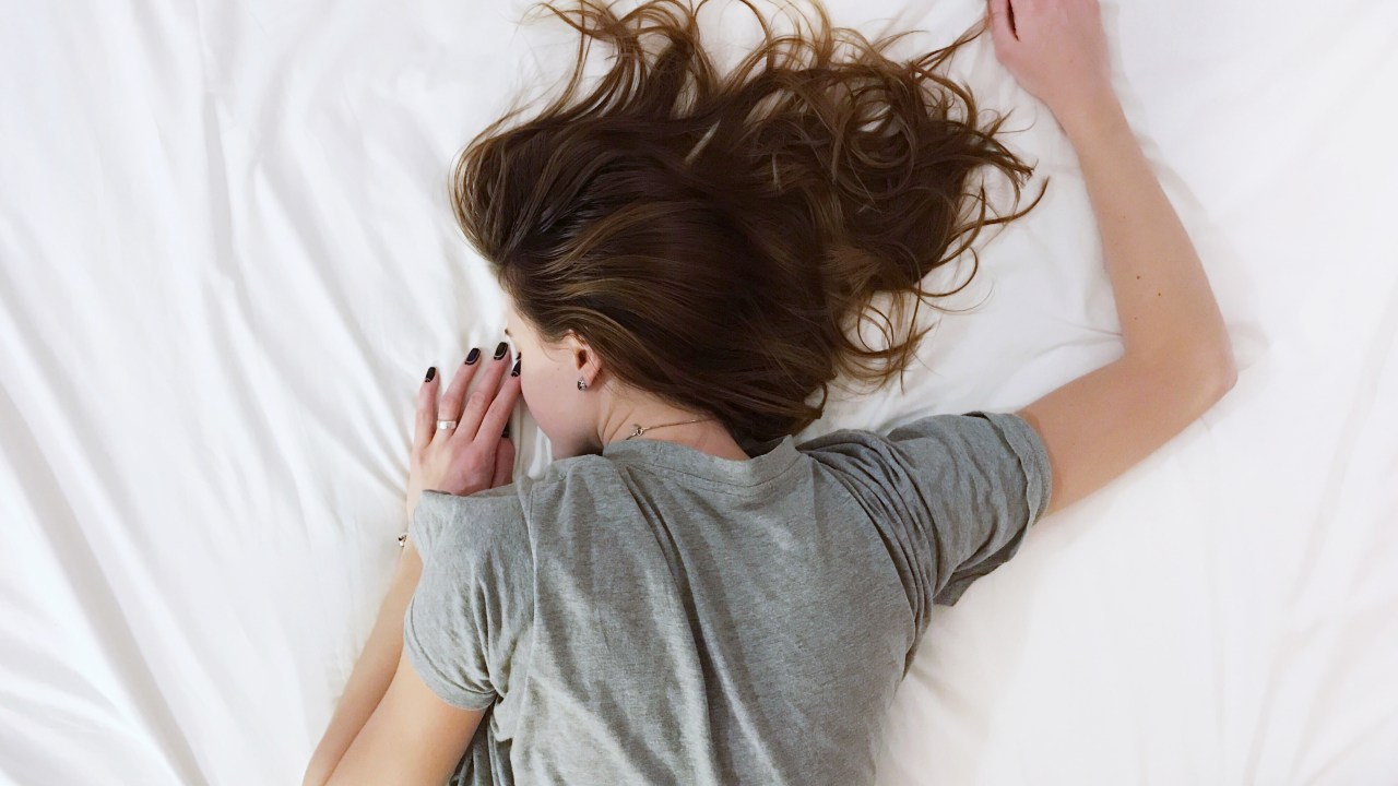 garota de camiseta cinza dormindo de bruços em cama grande e branca