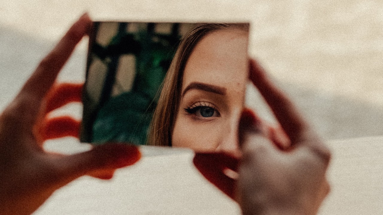 mulher olhando parte de seu reflexo (olho e sobrancelha) por espelho de mão
