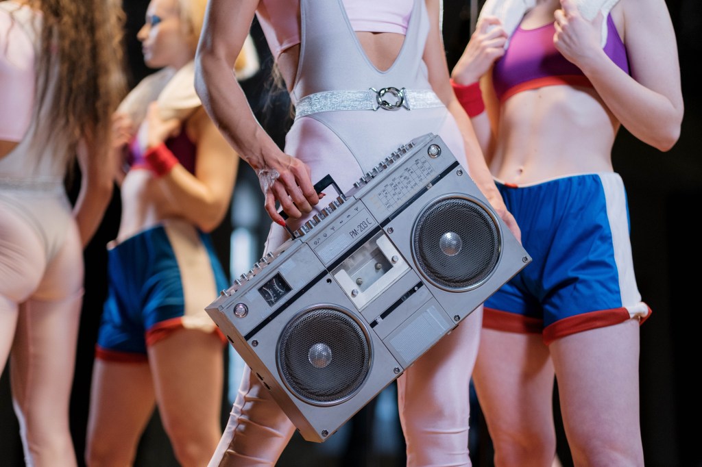 mulheres com roupa de academia inspirada nos anos 80 com uma segurando uma caixa de som