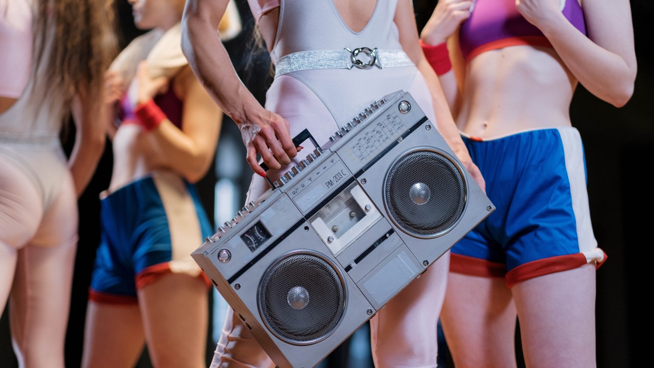 mulheres com roupa de academia inspirada nos anos 80 com uma segurando uma caixa de som