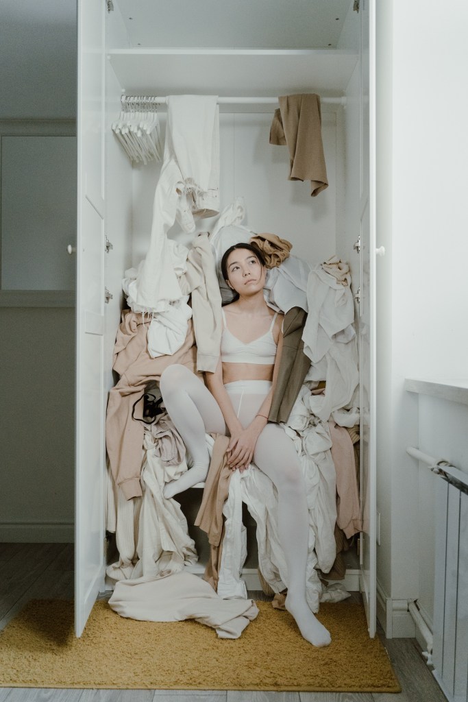 Mulher sentada em um armário com varias roupas caídas sobre ela. Sua expressão é pensativa, enquanto olha para cima.