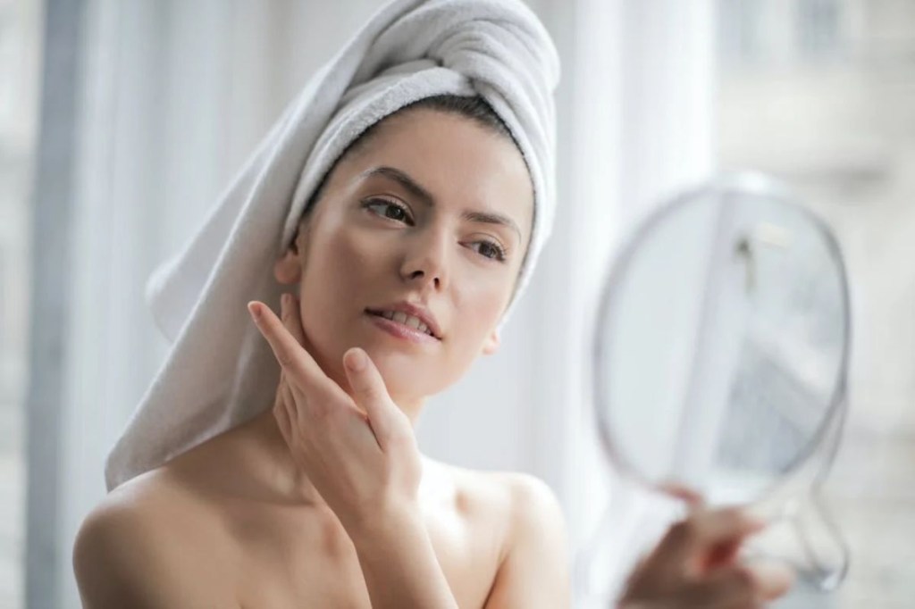 Mulher se olhando no espelho com toalha no cabelo e mão no rosto