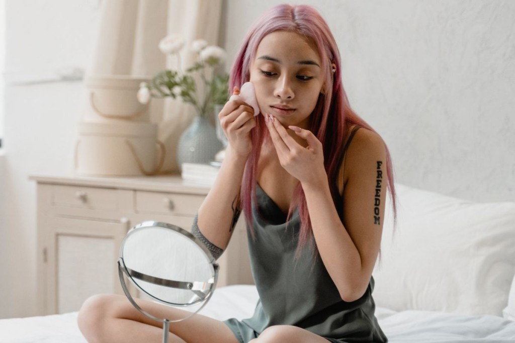 massageadores-faciais (1) Mulher massageando o rosto com Gua Sha em frente ao espelho