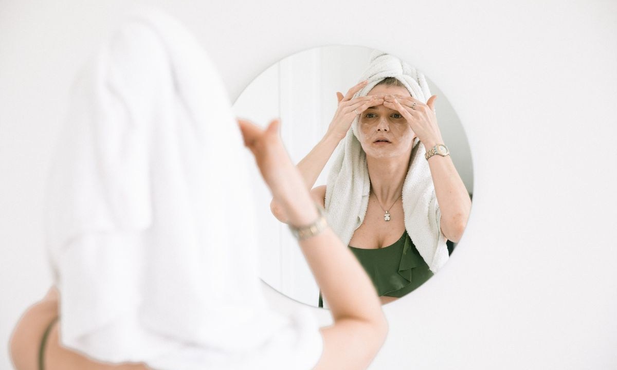 Mulher com toalha na cabeça passando produtos na pele em frente ao espelho