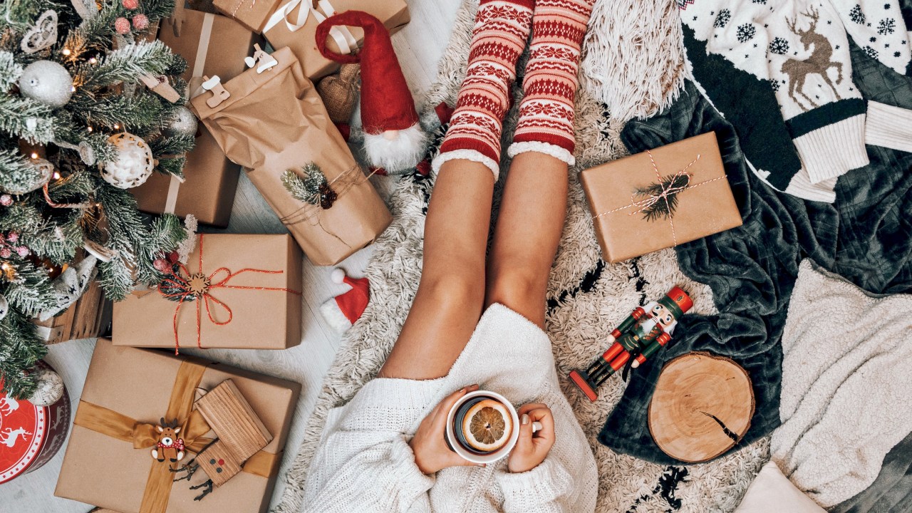 Mulher sentada de pijamas no chão tomando um chocolate quente e cercada de decoração de Natal