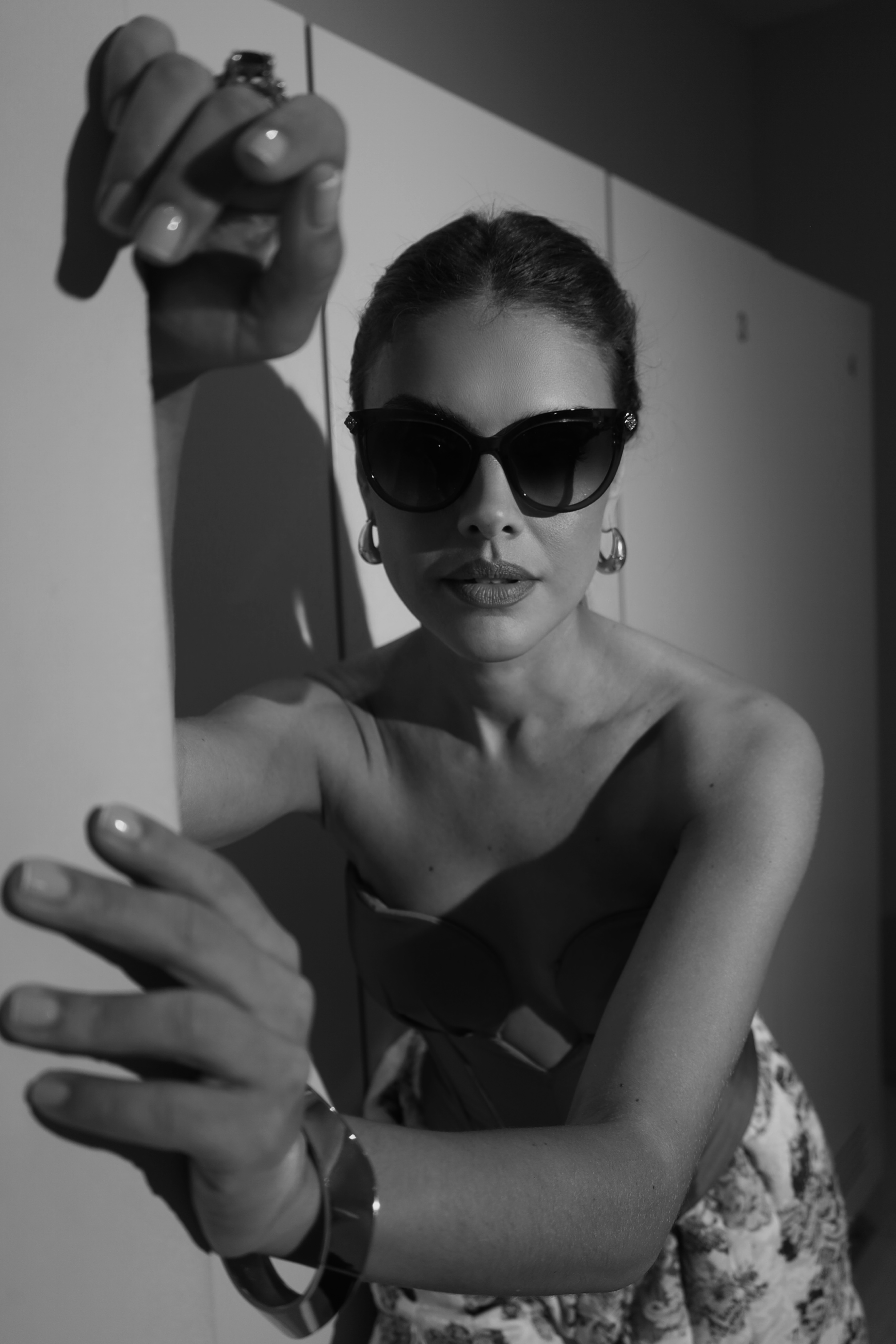 Mulher com &oacute;culos escuros e batom, cabelo preso, blusa tomara que caia e saia estampada, apoiada em uma parede clara, com luz e sombra marcantes