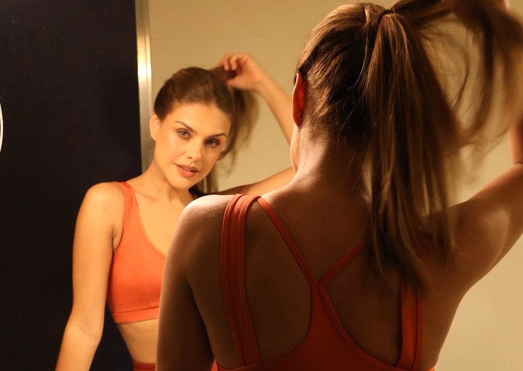 Mulher de costas, com cabelo castanho preso em rabo de cavalo, vestindo top esportivo laranja, olhando seu reflexo no espelho. No reflexo, ela sorri levemente e ajeita o cabelo