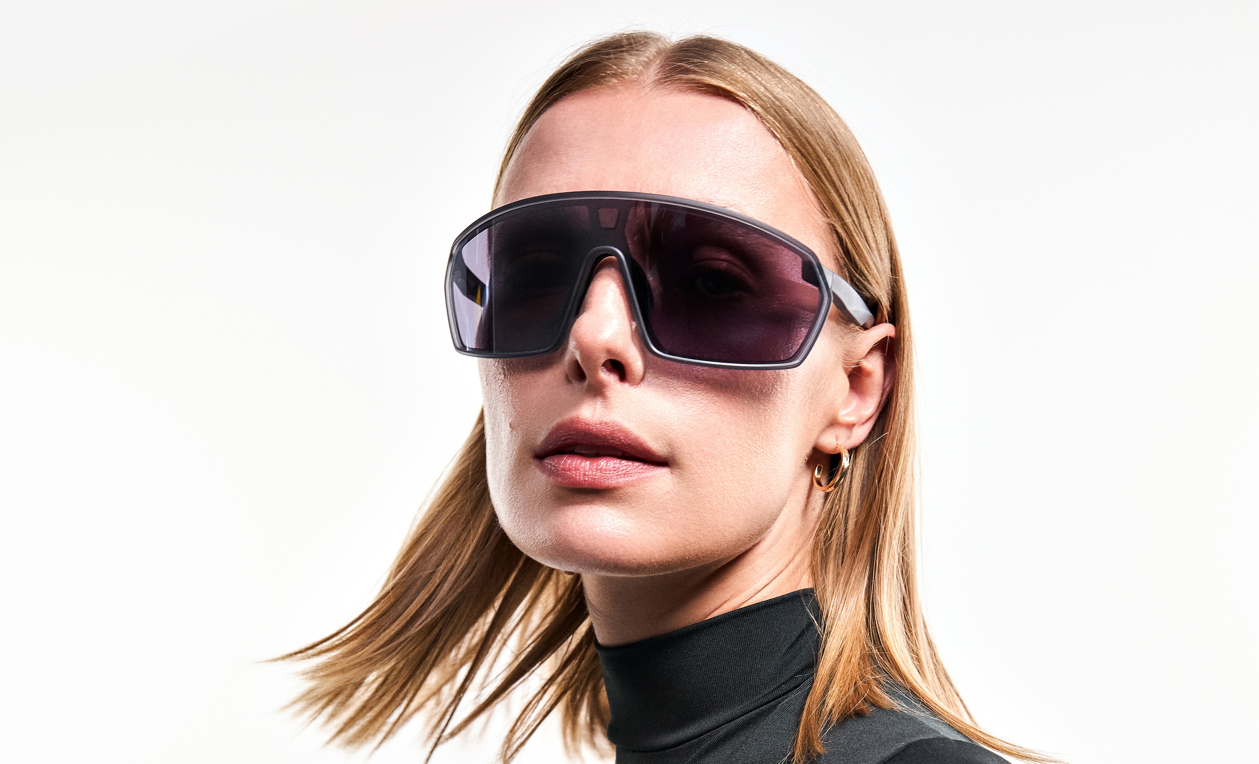 Mulher loira de óculos escuros esportivos com lentes roxas, vestindo blusa preta de gola alta e brinco de argola dourado, olhando para frente com expressão séria em fundo branco