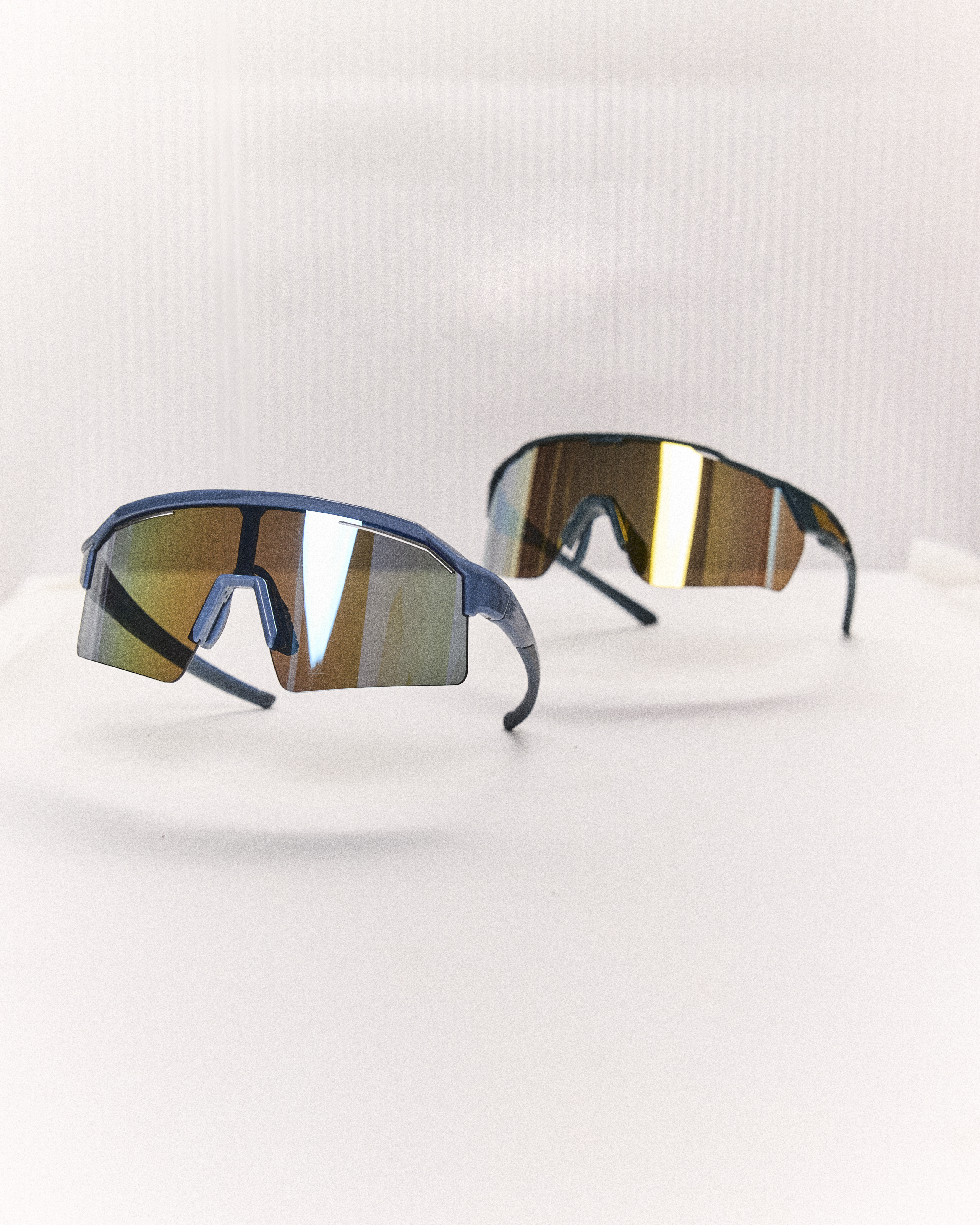 Dois óculos esportivos com lentes espelhadas coloridas, um azul escuro em primeiro plano e outro preto desfocado ao fundo, sobre uma superfície branca