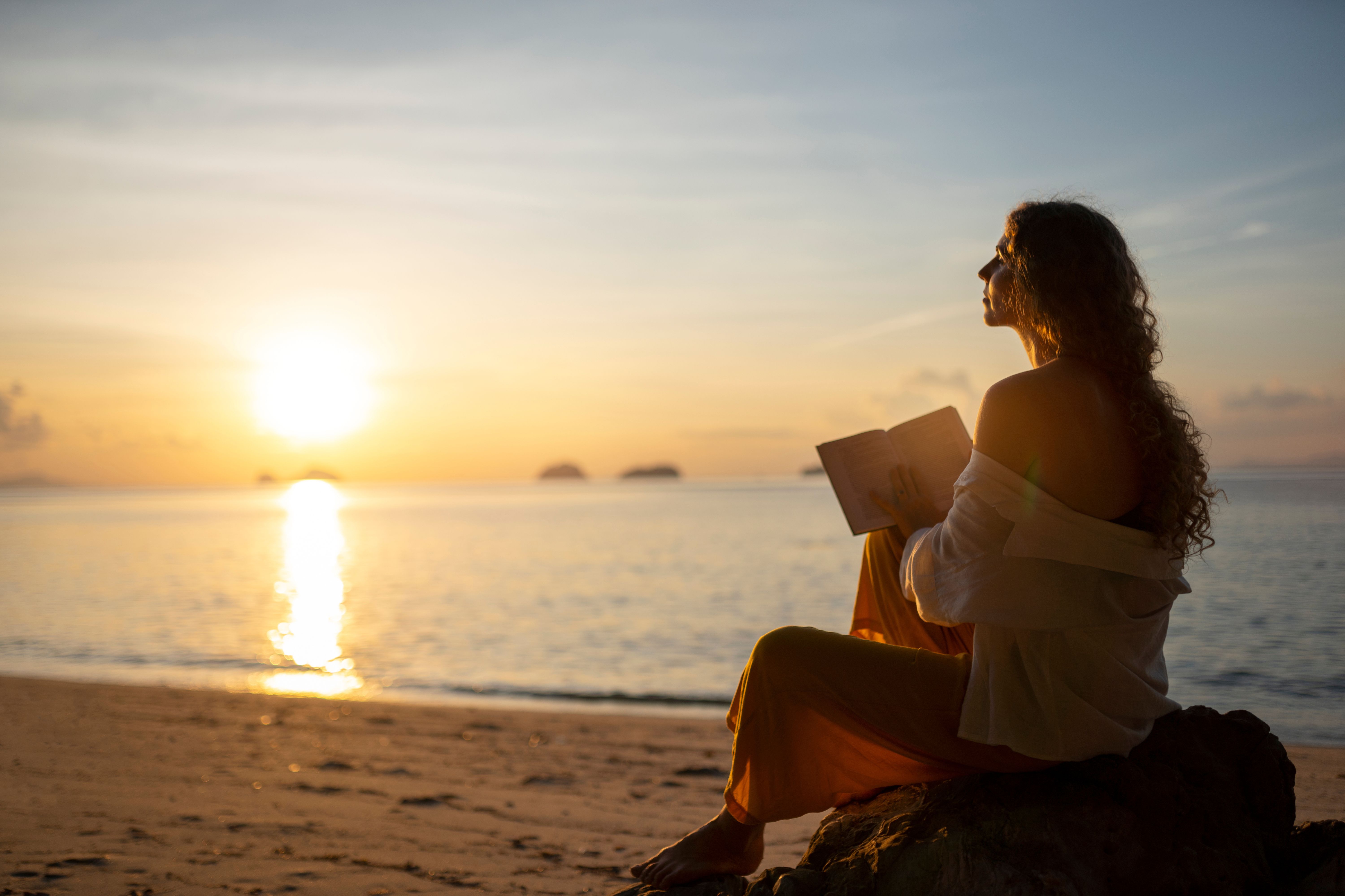 Mulher de cabelos cacheados, vestindo calça laranja e camisa branca, sentada em uma rocha na praia, lendo um livro ao pôr do sol. O sol brilha no horizonte, refletindo na água calma do mar, com pequenas ilhas ao fundo
