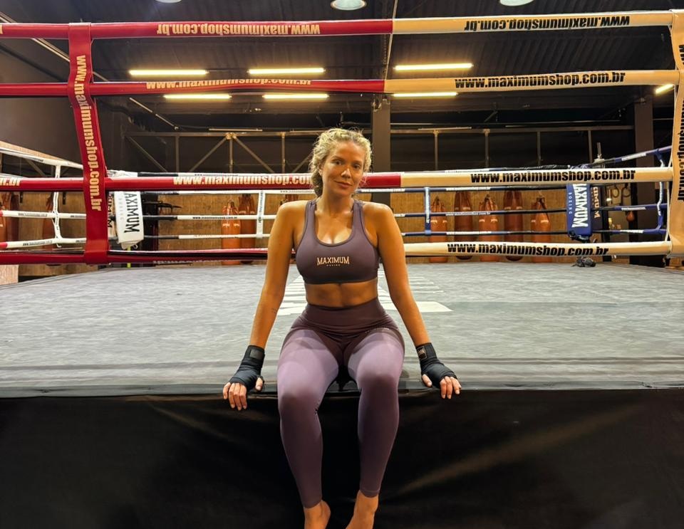 Mulher loira, de top e calça legging roxos, sentada descalça na beirada de um ringue de boxe, com faixas pretas nas mãos. O ringue tem cordas vermelhas e brancas com o site "www.maximumshop.com.br" repetido. Ao fundo, sacos de pancada marrons.