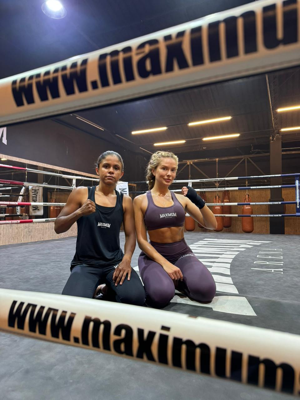Duas mulheres, uma morena e uma loira, ajoelhadas em um ringue de boxe, vestindo roupas esportivas da marca Maximum. A morena usa regata e calça preta, punho cerrado. A loira usa top e calça roxa, com bandagens nas mãos. Ambas olham para frente, com o nome da marca visível nas cordas do ringue e ao fundo. Sacos de pancada pendurados completam o cenário da academia