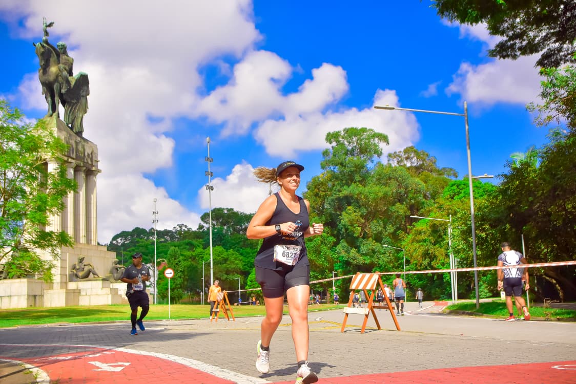 Mulher sorridente correndo em uma prova de rua, vestindo regata preta, shorts e boné, com o número 6827. Ao fundo, um monumento equestre, árvores e céu azul com nuvens brancas. Outros corredores e sinalização de corrida são visíveis na pista.