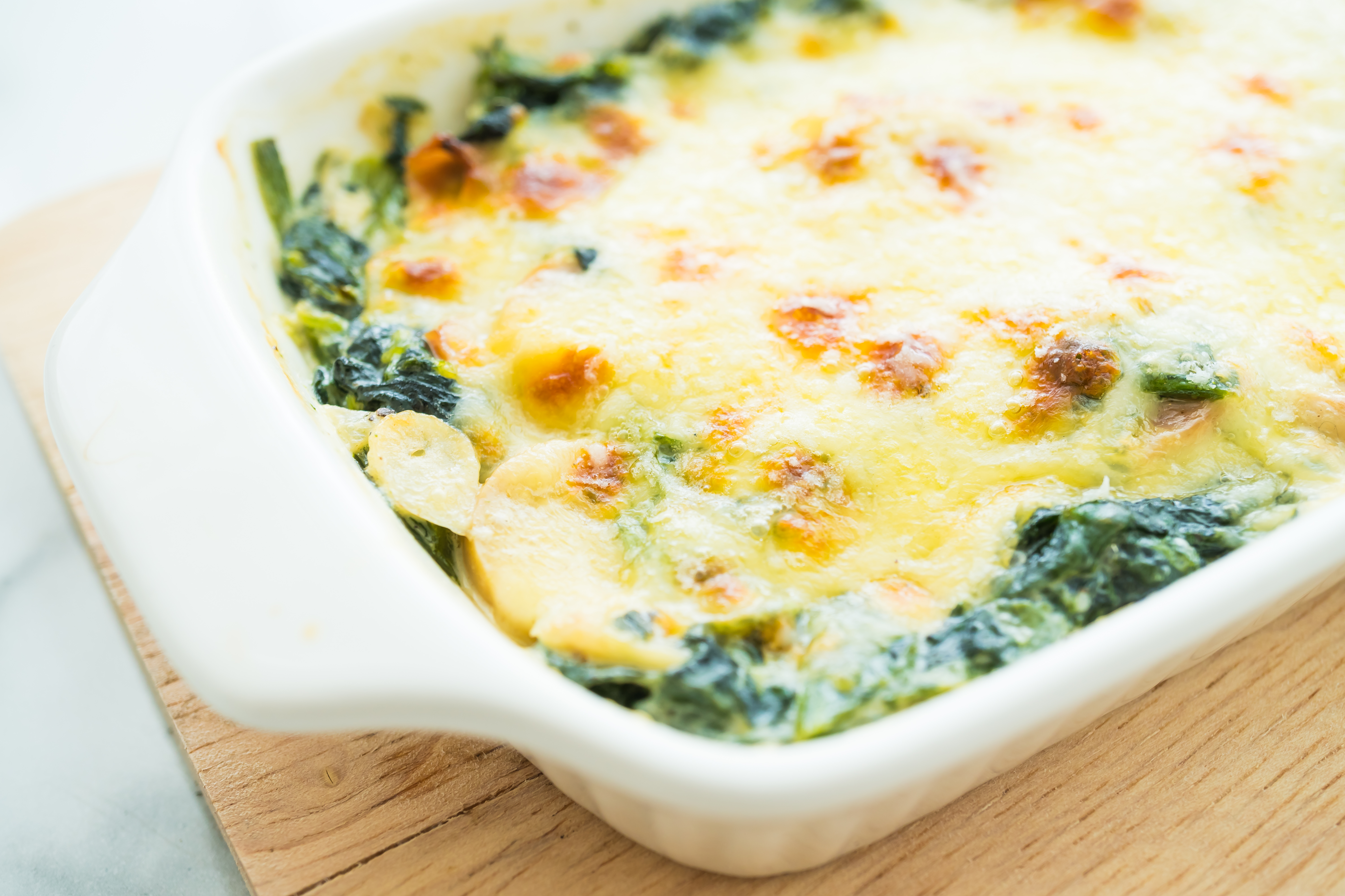 Assadeira branca com gratinado de espinafre e batata, coberto por queijo dourado e borbulhante, sobre uma tábua de madeira