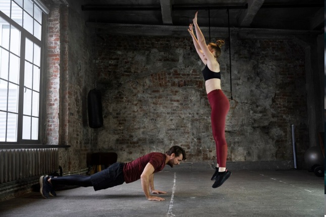 Homem fazendo flexão no chão e mulher pulando com os braços erguidos em uma academia com paredes de tijolos e janela grande