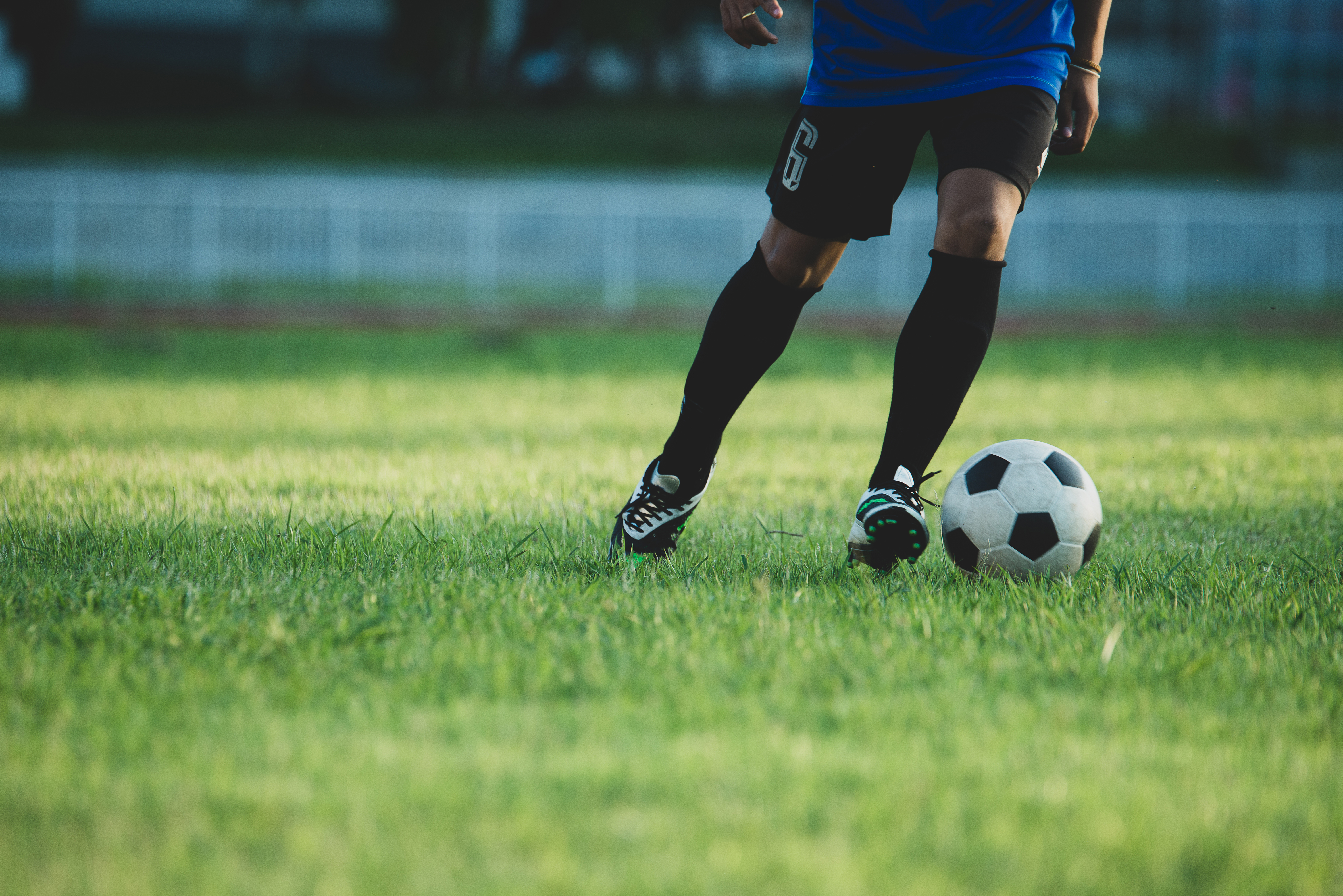Pernas de um jogador de futebol com uniforme azul e preto, meias e chuteiras pretas, driblando uma bola branca e preta em um campo de grama verde