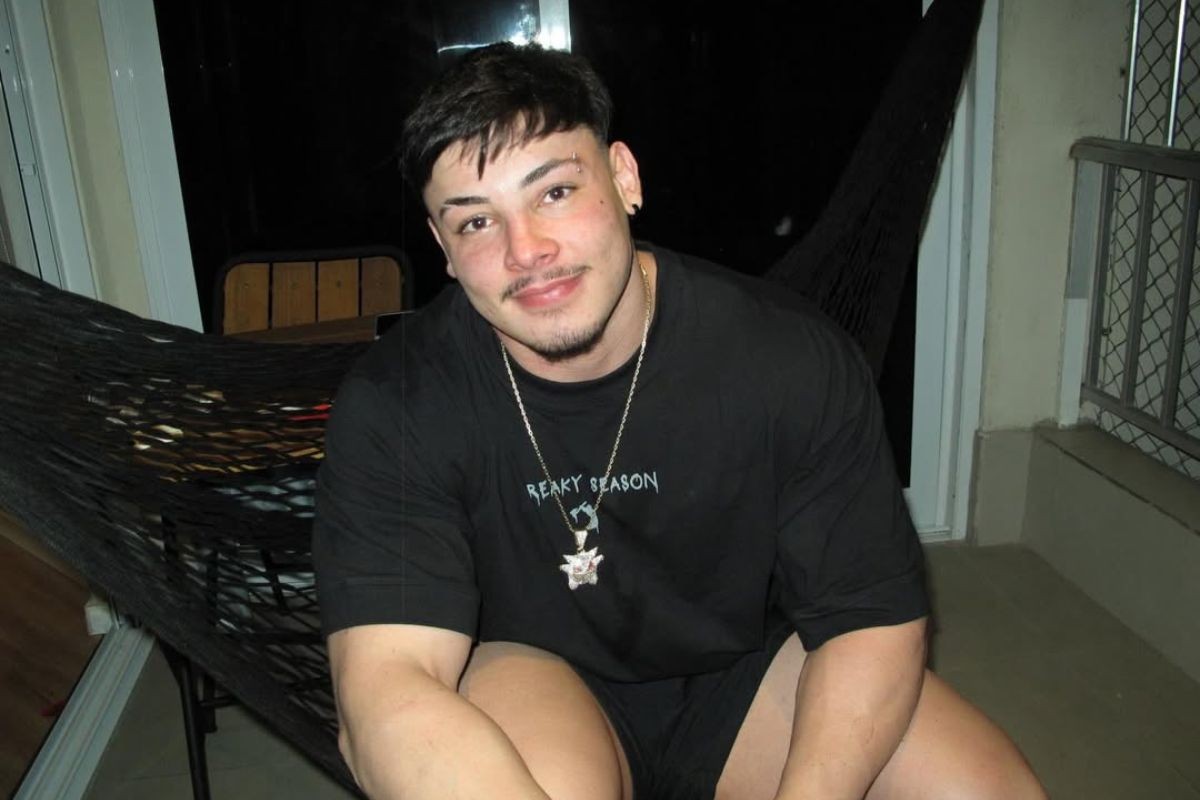 Homem jovem com bigode e cavanhaque, usando camiseta preta e shorts, sentado em uma rede escura em um ambiente noturno, sorrindo para a câmera