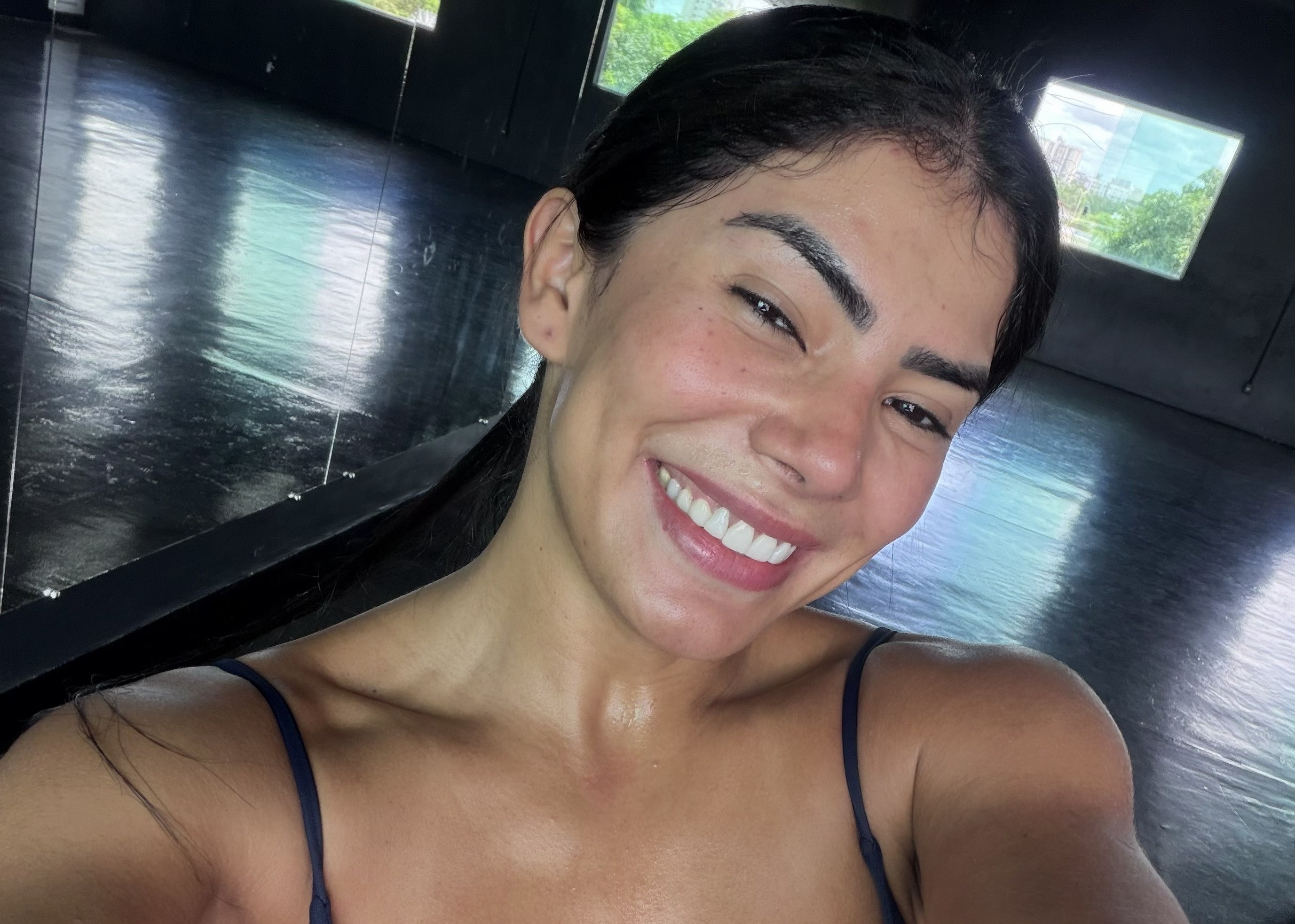 Mulher de pele morena clara, cabelo escuro preso, sorrindo com dentes brancos e maquiagem leve, vestindo top azul escuro. O fundo é um ambiente interno com chão escuro e reflexivo, e uma janela retangular mostrando folhagem verde e céu claro