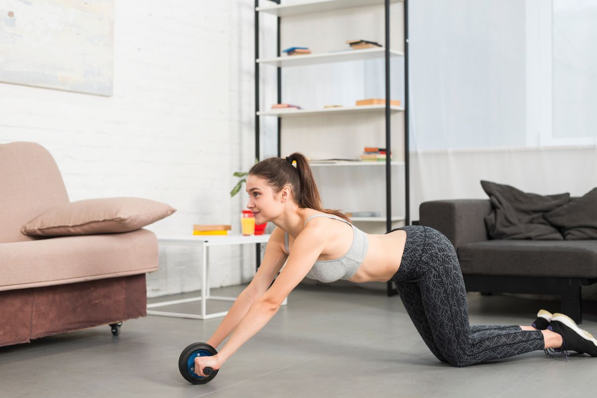 Mulher jovem de cabelo castanho preso, usando top esportivo cinza e calça legging estampada, fazendo exercício abdominal com um rolo de fitness no chão da sala, com um sofá e estante ao fundo