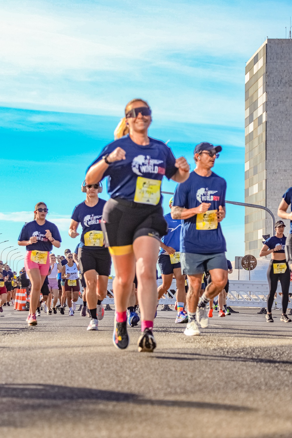 Corredores participam de uma maratona em rua asfaltada, sob c&eacute;u azul claro. A mulher em primeiro plano, sorrindo, veste camiseta azul 