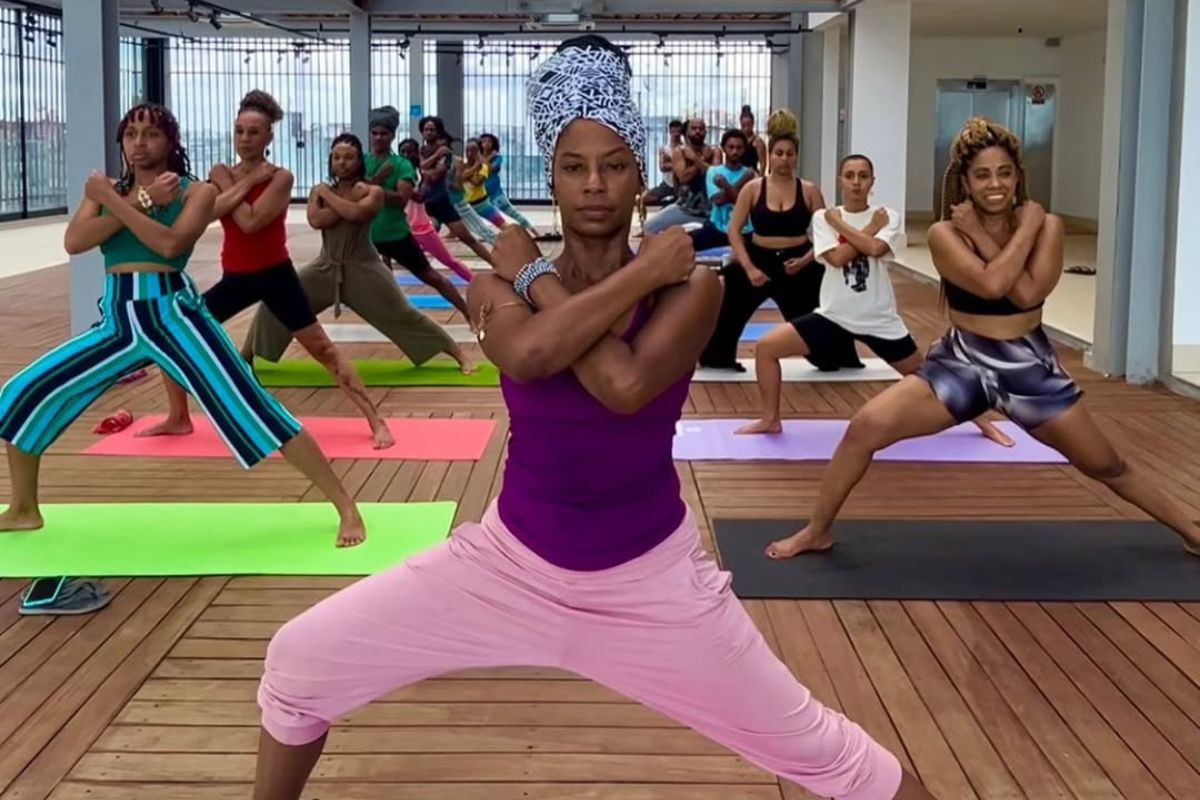 Mulheres negras praticando yoga em um estúdio com piso de madeira, usando roupas coloridas e fazendo a postura do guerreiro com braços cruzados no peito