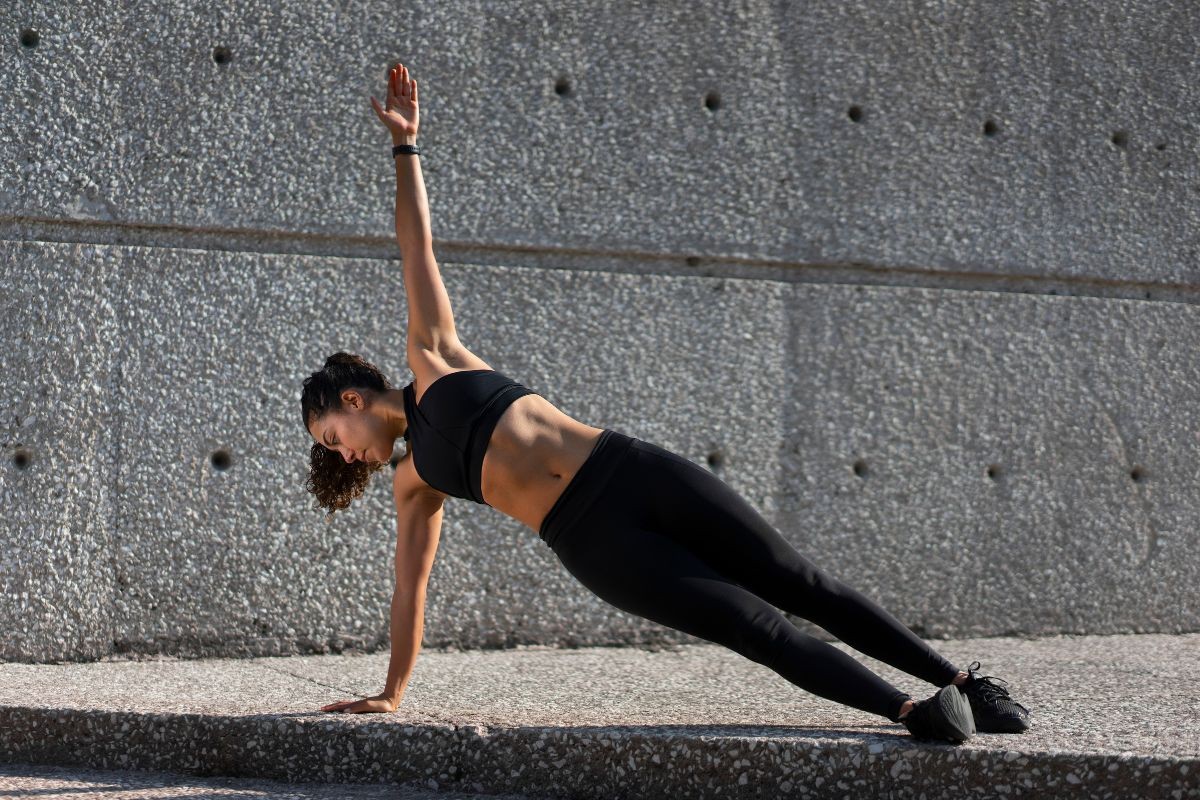 Mulher jovem de cabelo cacheado escuro, vestindo top e calça legging pretos, fazendo prancha lateral com o braço direito estendido para cima, em um muro de concreto cinza