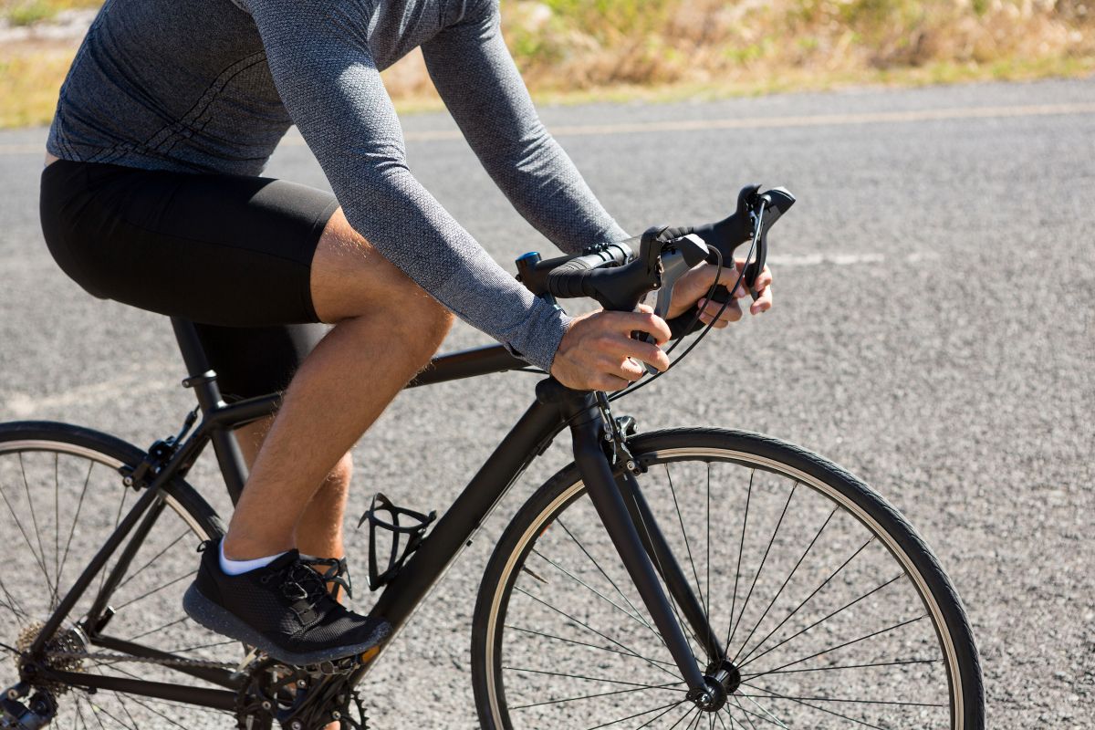 Ciclista masculino com camisa cinza de manga comprida e bermuda preta, pedalando uma bicicleta de estrada preta em uma pista asfaltada, com as mãos no guidão e os pés nos pedais