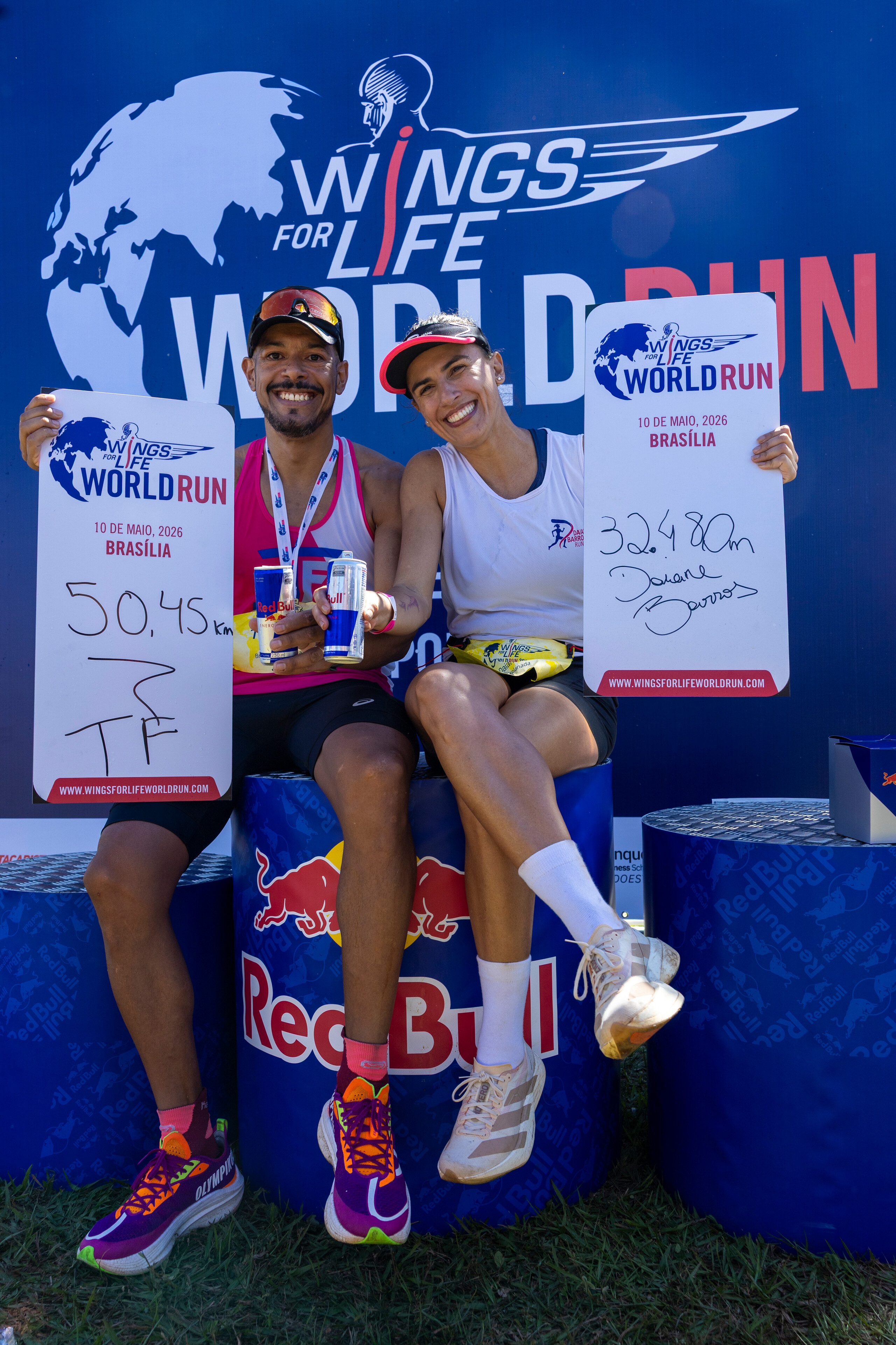 Dois corredores sorridentes, um homem e uma mulher, sentados em barris azuis da Red Bull, exibem placas brancas com seus resultados da Wings for Life World Run 2026 em Bras&iacute;lia. O homem, &agrave; esquerda, veste regata rosa e segura duas latas de Red Bull, sua placa mostra 