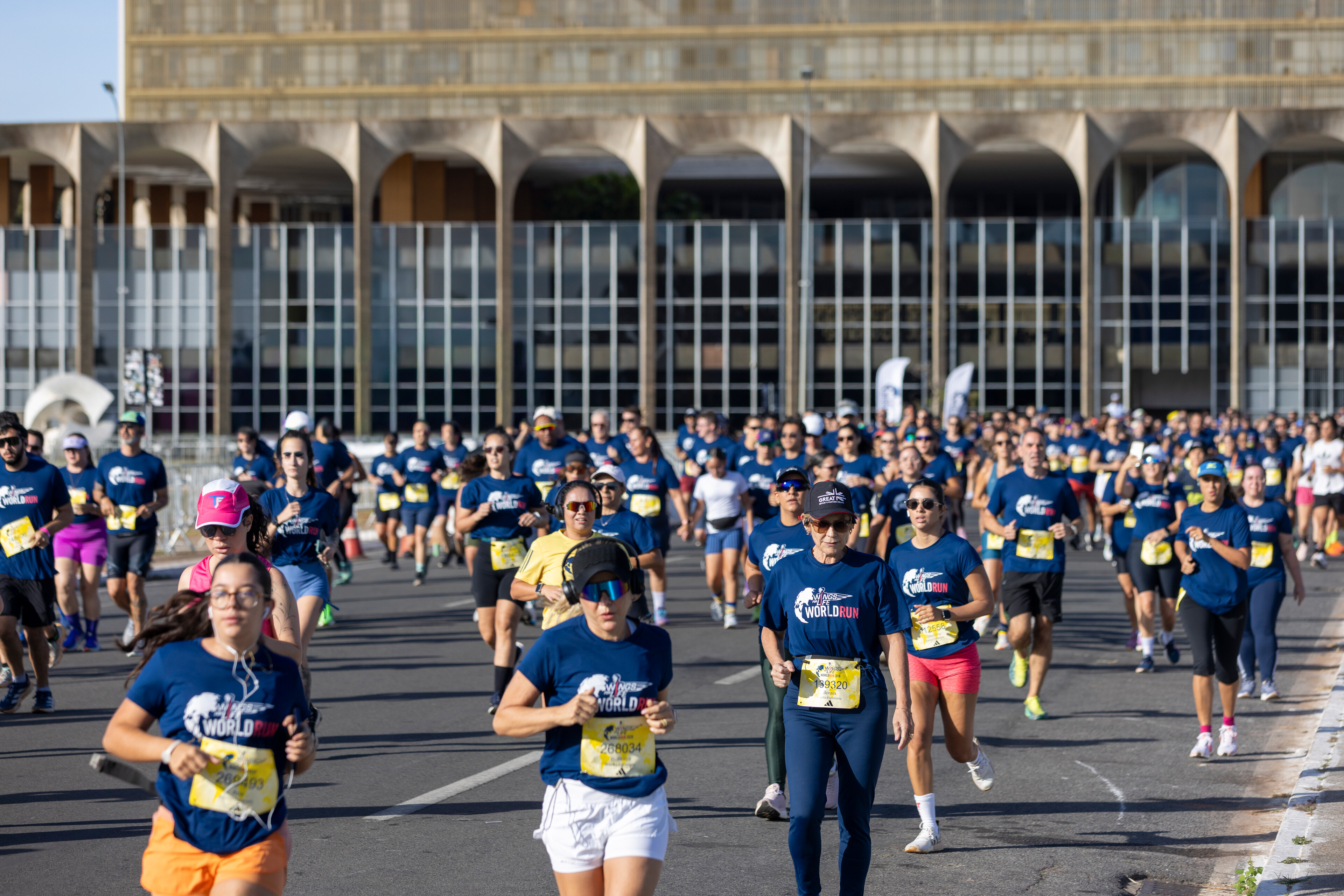 Multidão de corredores de diversas idades, vestindo camisetas azuis com "World Run", correndo em uma rua larga com um grande edifício de arquitetura moderna ao fundo