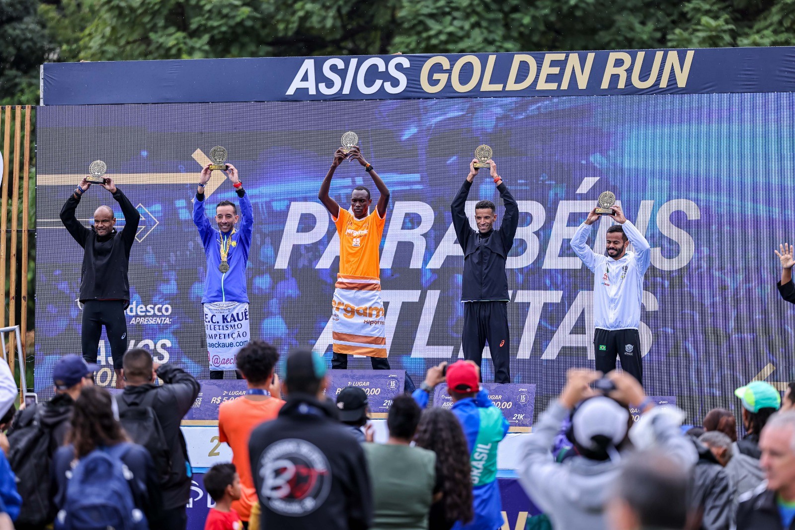 Cinco atletas masculinos sorridentes no pódio da Asics Golden Run, erguendo troféus. O vencedor, ao centro, veste laranja, os demais usam roupas esportivas escuras e claras. Uma multidão os observa e fotografa