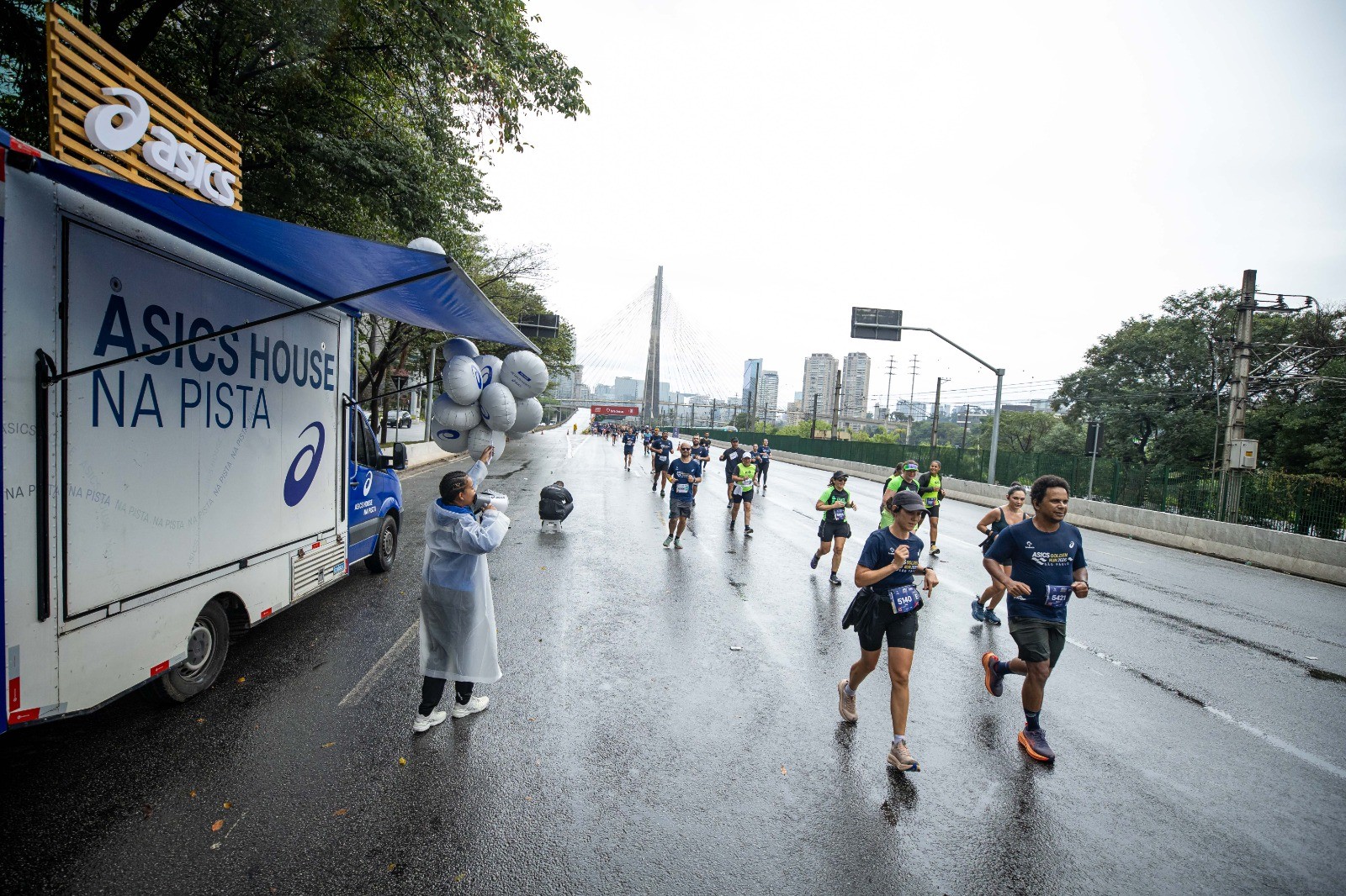 Corredores em uma maratona na rua molhada, com um caminhão branco da Asics à esquerda e o logotipo da marca no topo. Uma pessoa de capa de chuva segura balões brancos ao lado do caminhão. Ao fundo, prédios e a Ponte Estaiada de São Paulo sob céu nublado