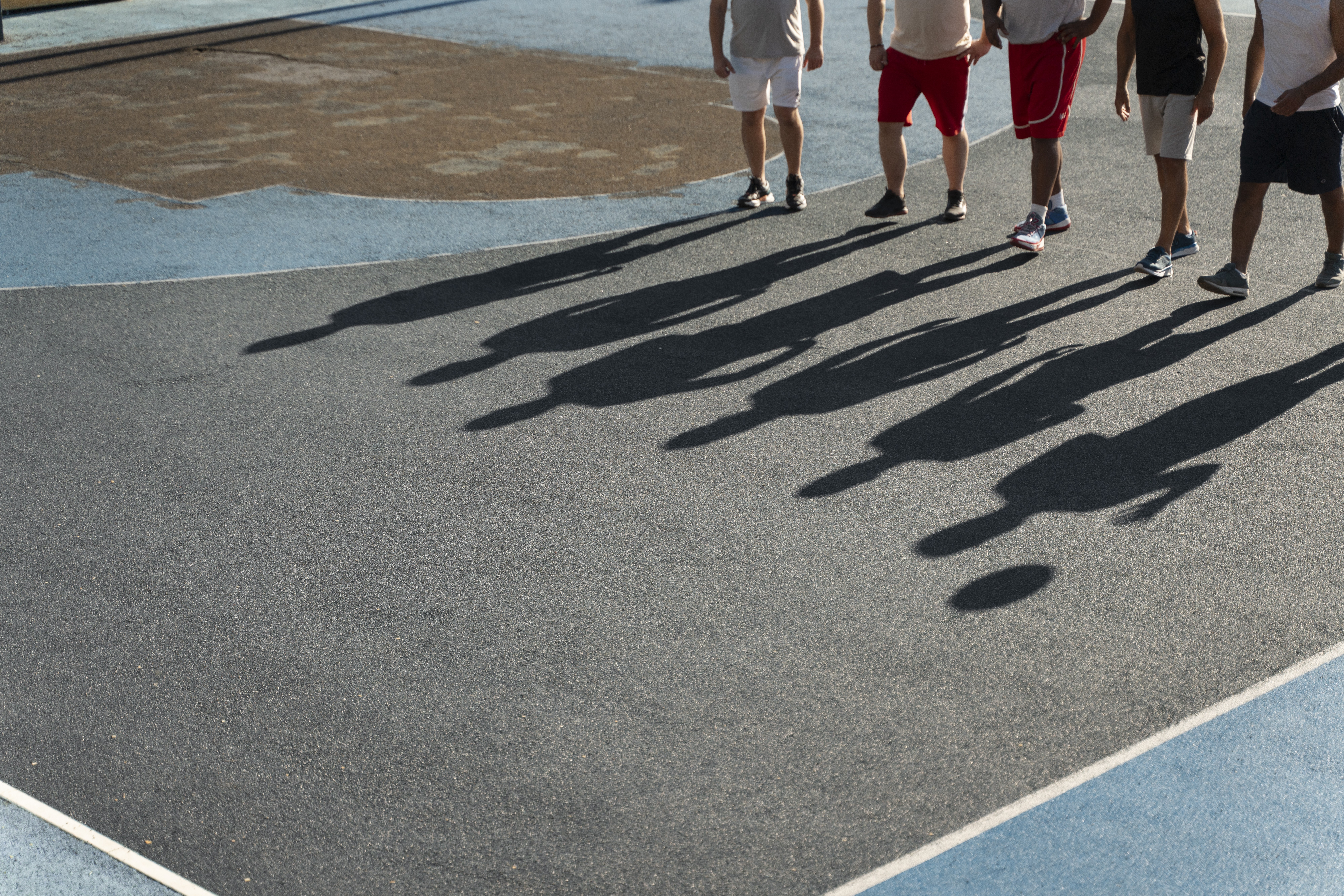 Cinco homens em shorts e tênis, com pernas visíveis, parados em uma quadra esportiva cinza com linhas azuis e marrons, projetando longas sombras escuras no chão