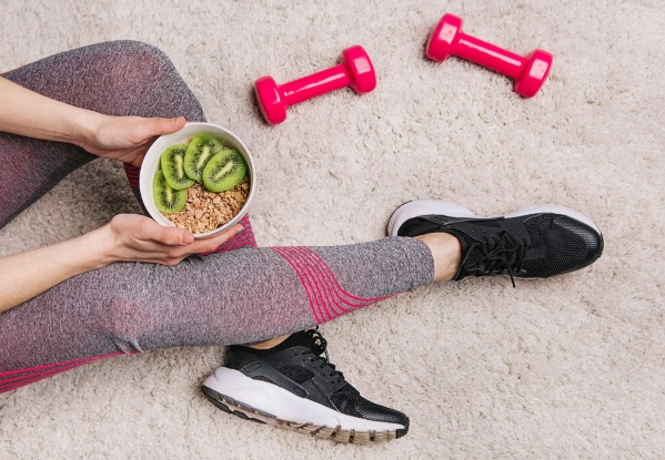Pessoa sentada no chão com leggings cinza e rosa, segurando uma tigela de granola com kiwi fatiado. Ao lado, dois halteres rosa e tênis esportivos pretos, sobre um tapete felpudo branco